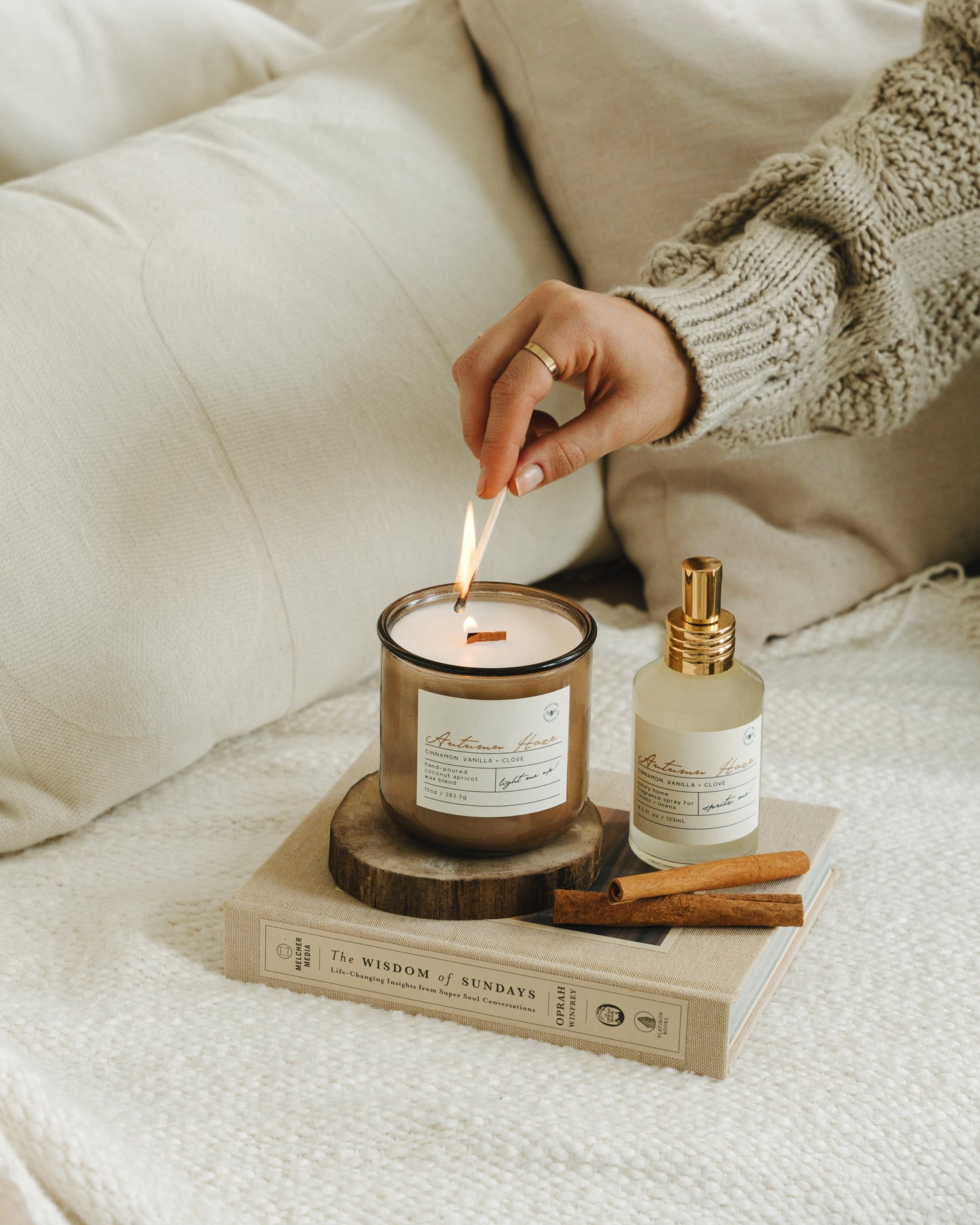 Person lighting a candle on a cozy sofa with a bottle of diffuser and cinnamon sticks.